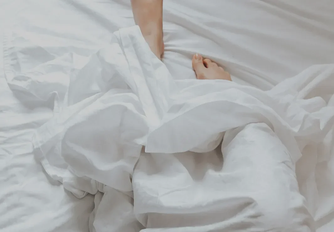 White sheets and duvet rumpled on a bed, with a bare foot and hand visible.